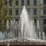 Strūklaka pilsētas centrā pie vēsturiskām ēkām. Redzama daudzlīmeņu strūklaka ar vairākiem ūdens strūklu lokiem un apkārtējo apstādījumu daļām. Rīgas strūklaku ieziemošana 2026 noslēgs aptuveni sešus mēnešus ilgo strūklaku sezonu.