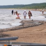 Rīgas peldvietā ar tīru ūdeni un atpūtniekiem pie ūdens, ilustrējot labu ūdens kvalitāti, kas atbilst higiēnas prasībām visās oficiālajās un populārajās peldvietās pilsētā. Vecāķu pludmale.