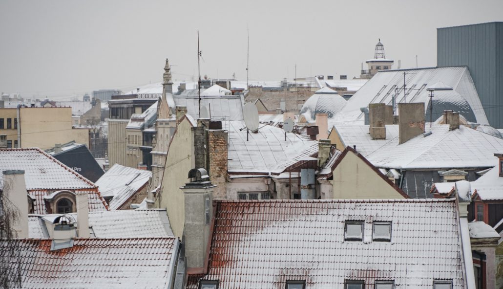 Dzīvojamā ēka Rīgā atkušņa laikā, uz jumta redzams kūstošs sniegs un lāstekas, uzsverot nepieciešamību regulāri tīrīt jumtu un uzturēt notekcaurules, lai novērstu bīstamas situācijas gājējiem un autovadītājiem.