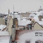 Dzīvojamā ēka Rīgā atkušņa laikā, uz jumta redzams kūstošs sniegs un lāstekas, uzsverot nepieciešamību regulāri tīrīt jumtu un uzturēt notekcaurules, lai novērstu bīstamas situācijas gājējiem un autovadītājiem.