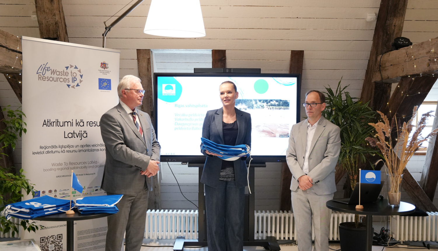 Zilais karogs Daugavgrīvas peldvietā Rīgā, tīra pludmale ar sakoptu infrastruktūru, kas atbilst starptautiskajiem vides kvalitātes standartiem un nodrošina drošu atpūtu pie ūdens.