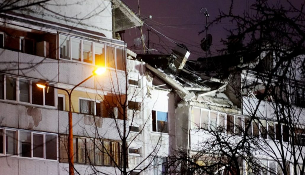 Bauskas ielas 15 daudzdzīvokļu ēka pēc sprādziena Rīgā. Attēlā redzami nopietni jumta un ēkas konstrukciju bojājumi, bojāta fasāde, apgaismoti logi, ielas apgaismojums un koku zari priekšplānā nakts laikā. Attēls ilustrē Bauskas ielas sprādzienu, Rīgas pašvaldības atbalstu un mājokļu atjaunošanu cietušajiem iedzīvotājiem.