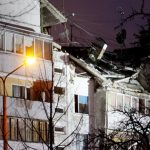Bauskas ielas 15 daudzdzīvokļu ēka pēc sprādziena Rīgā. Attēlā redzami nopietni jumta un ēkas konstrukciju bojājumi, bojāta fasāde, apgaismoti logi, ielas apgaismojums un koku zari priekšplānā nakts laikā. Attēls ilustrē Bauskas ielas sprādzienu, Rīgas pašvaldības atbalstu un mājokļu atjaunošanu cietušajiem iedzīvotājiem.