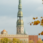 Rīgas pašvaldība informē par gaisa kvalitāti: redzama Rīgas panorāma rudenī ar baznīcas torni, ēku jumtiem un dzeltenīgām koku lapām. Fonā redzamas mākoņainas debesis virs pilsētvides.