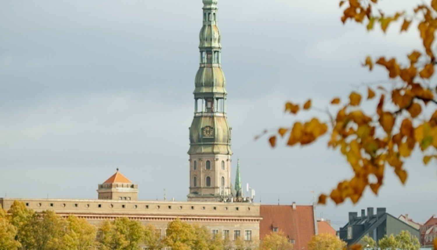 Rīgas pašvaldība informē par gaisa kvalitāti: redzama Rīgas panorāma rudenī ar baznīcas torni, ēku jumtiem un dzeltenīgām koku lapām. Fonā redzamas mākoņainas debesis virs pilsētvides.