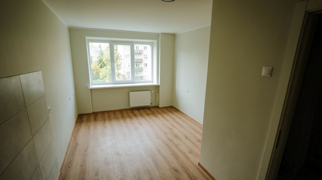 Atjaunots sociālais dzīvoklis Rīgā ar gaišām sienām, lamināta grīdu, logu un radiatoru istabas galā. Attēlā redzama dzīvošanai sagatavota telpa pēc remonta. Rīgas sociālo mājokļu atjaunošana nodrošina kvalitatīvu dzīves vidi iedzīvotājiem.