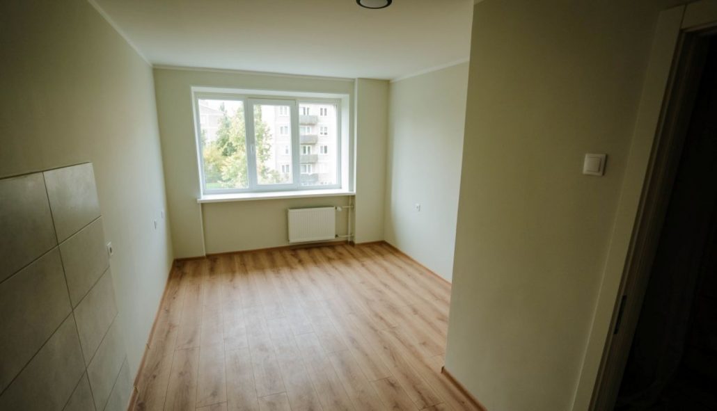 Atjaunots sociālais dzīvoklis Rīgā ar gaišām sienām, lamināta grīdu, logu un radiatoru istabas galā. Attēlā redzama dzīvošanai sagatavota telpa pēc remonta. Rīgas sociālo mājokļu atjaunošana nodrošina kvalitatīvu dzīves vidi iedzīvotājiem.
