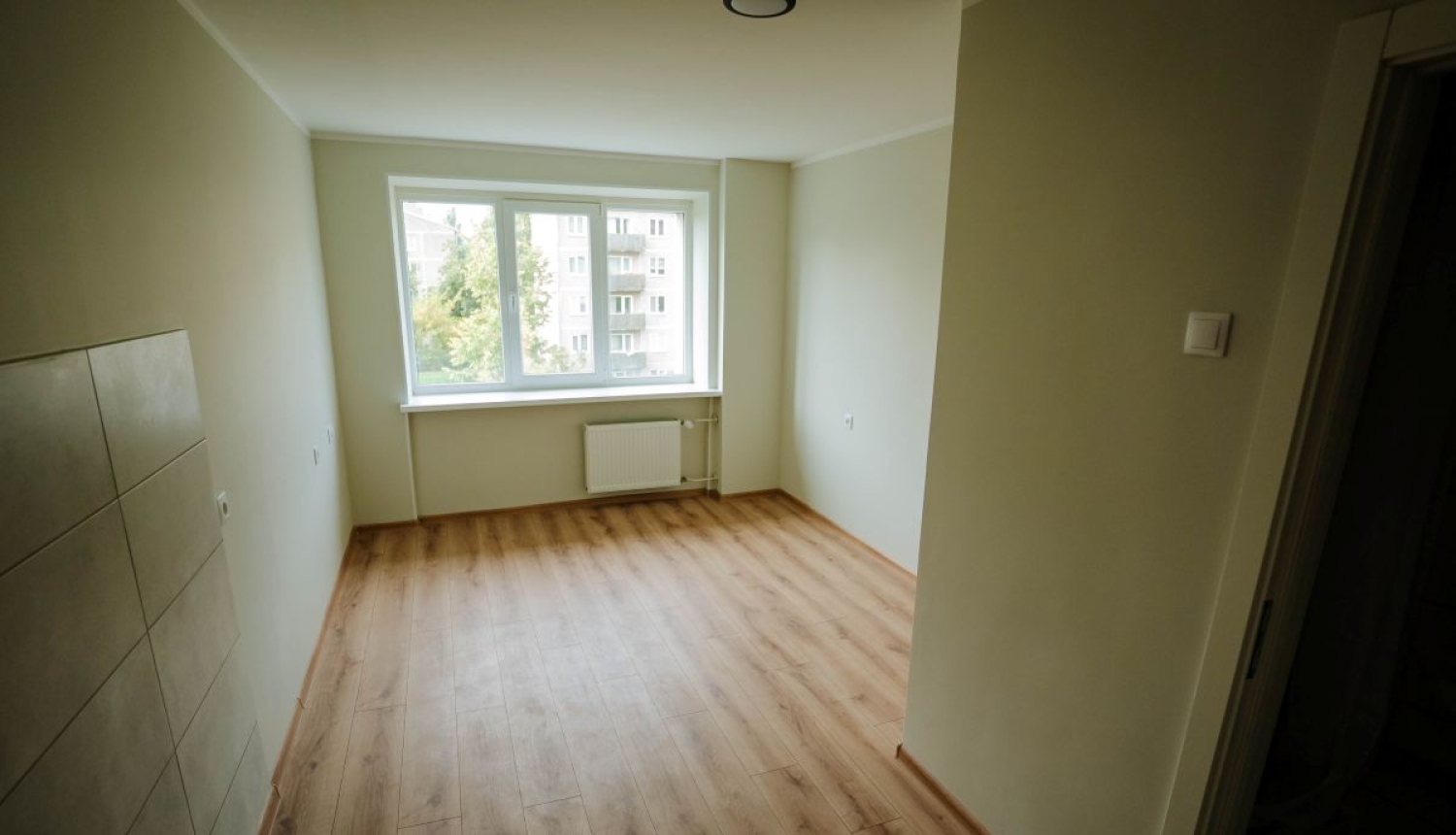 Atjaunots sociālais dzīvoklis Rīgā ar gaišām sienām, lamināta grīdu, logu un radiatoru istabas galā. Attēlā redzama dzīvošanai sagatavota telpa pēc remonta. Rīgas sociālo mājokļu atjaunošana nodrošina kvalitatīvu dzīves vidi iedzīvotājiem.