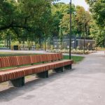 Rīgas parku dārzu apsaimniekošanas iepirkums: labiekārtots pilsētas parks ar garu koka soliņu gar pastaigu celiņu. Apkārt ir zāliens, koki un apstādījumi, fonā redzama parka infrastruktūra un sakopta publiskā ārtelpa.