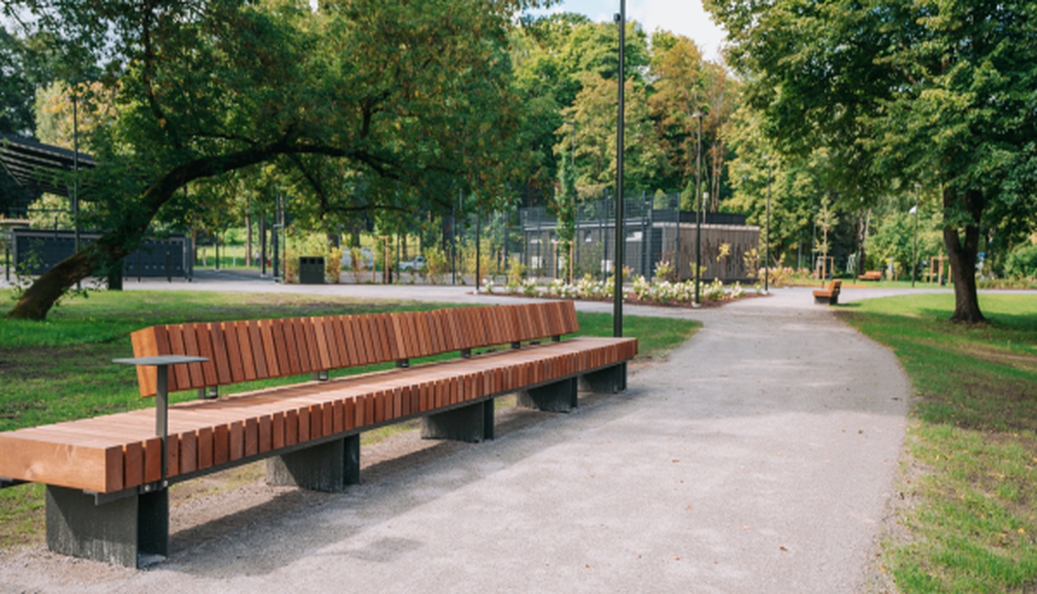 Rīgas parku dārzu apsaimniekošanas iepirkums: labiekārtots pilsētas parks ar garu koka soliņu gar pastaigu celiņu. Apkārt ir zāliens, koki un apstādījumi, fonā redzama parka infrastruktūra un sakopta publiskā ārtelpa.