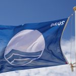 Rīgas Zilā karoga peldvietas: Zilā karoga karogs, kas plīvo vējā uz balta karoga masta. Karogs ir zilā krāsā ar baltu Zilā karoga simbolu centrā. Fonā redzamas zilas debesis un balti mākoņi.