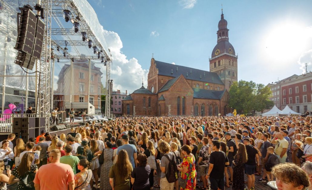 Liels publisks pasākums pilsētvidē ar koncertskatuvi un apmeklētāju pulcēšanos. Foto ilustrē vidi, kurā atkārtoti lietojamu trauku depozīta sistēmas var palīdzēt samazināt vienreizlietojamo iepakojumu un atkritumu daudzumu.