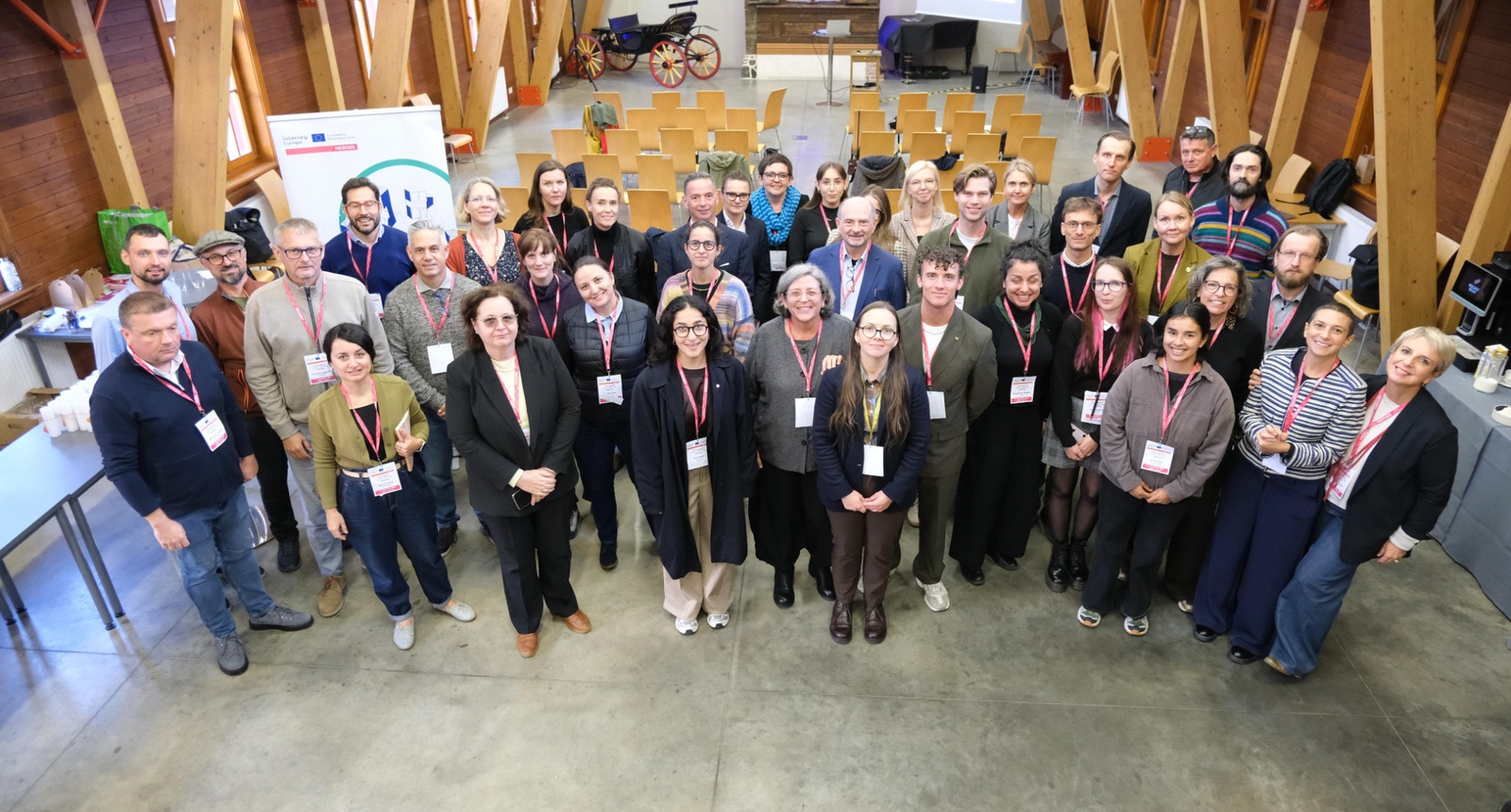 Starptautiska semināra dalībnieki grupas foto telpā ar koka konstrukcijām un prezentācijas aprīkojumu. Redzama projekta HEROES partneru un ekspertu grupa. Pieejamu mājokļu attīstība Rīgā bija galvenā pasākuma tēma.