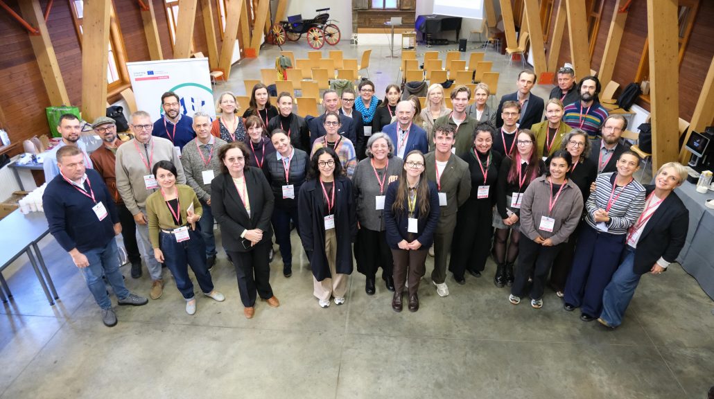 Starptautiska semināra dalībnieki grupas foto telpā ar koka konstrukcijām un prezentācijas aprīkojumu. Redzama projekta HEROES partneru un ekspertu grupa. Pieejamu mājokļu attīstība Rīgā bija galvenā pasākuma tēma.
