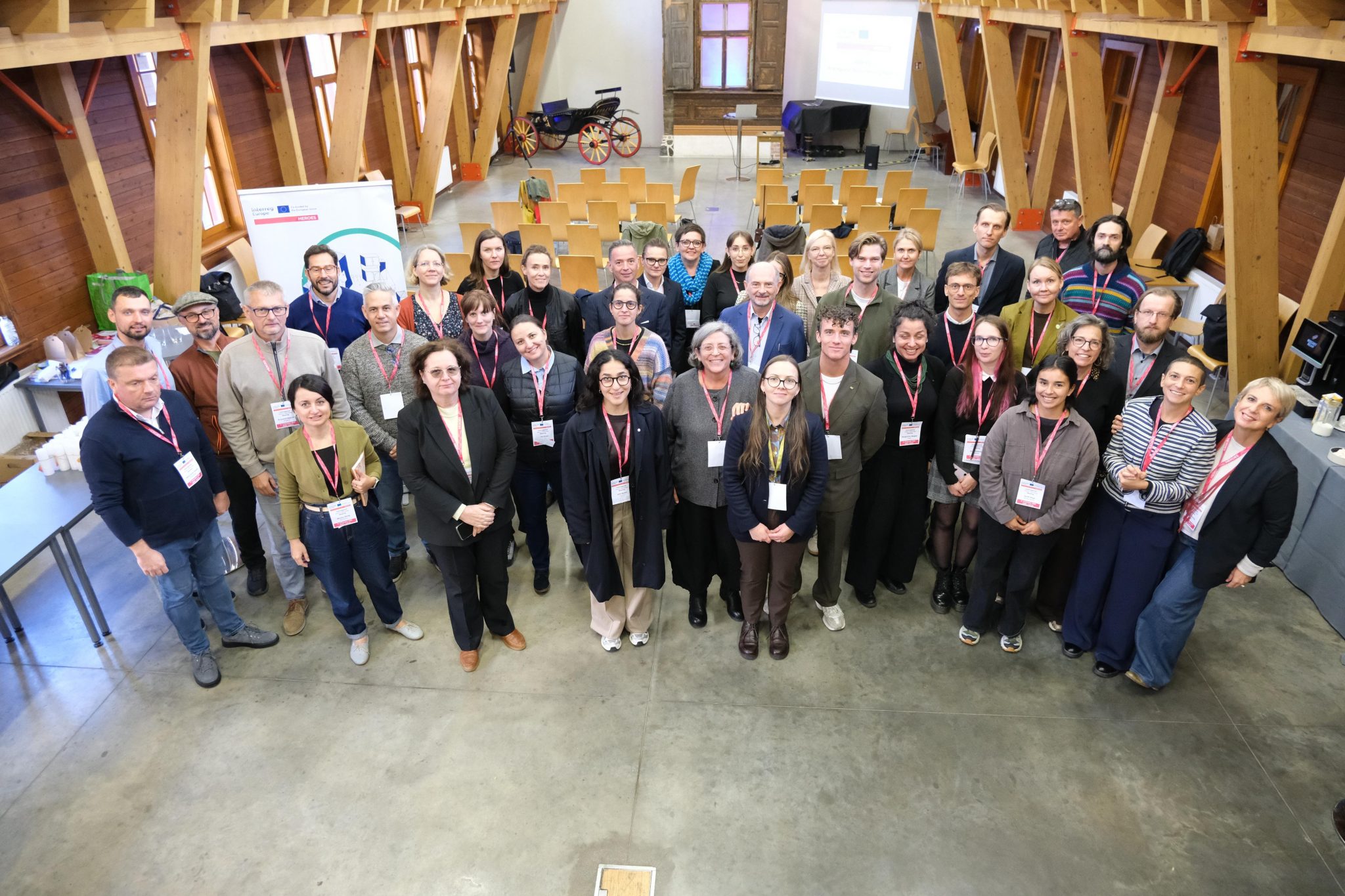 Starptautiska semināra dalībnieki grupas foto telpā ar koka konstrukcijām un prezentācijas aprīkojumu. Redzama projekta HEROES partneru un ekspertu grupa. Pieejamu mājokļu attīstība Rīgā bija galvenā pasākuma tēma.