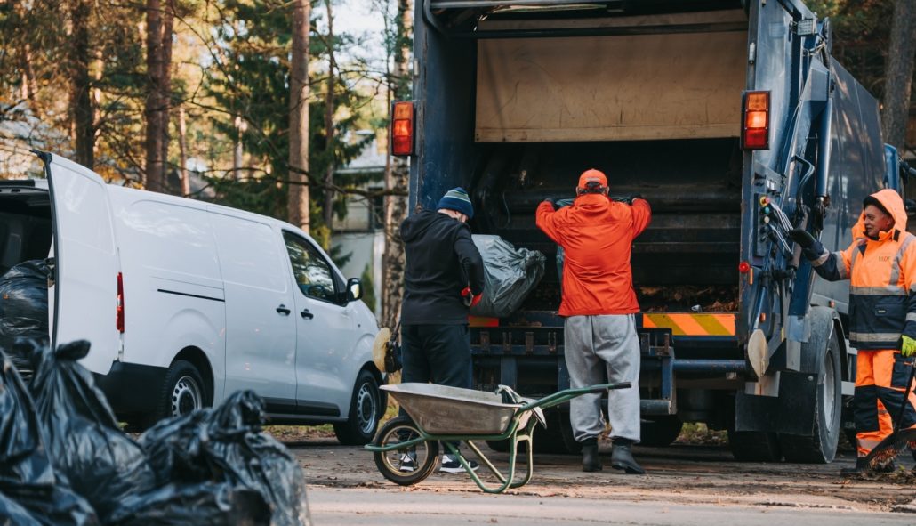 Iedzīvotāji pie specializētā atkritumu savākšanas transportlīdzekļa izkrauj savāktās rudens lapas. Attēlā redzami lapu maisi, ķerra un lapu savākšanas darbi. Attēls ilustrē rudens lapu pieņemšanas punktus Rīgā un lapu nodošanas procesu.